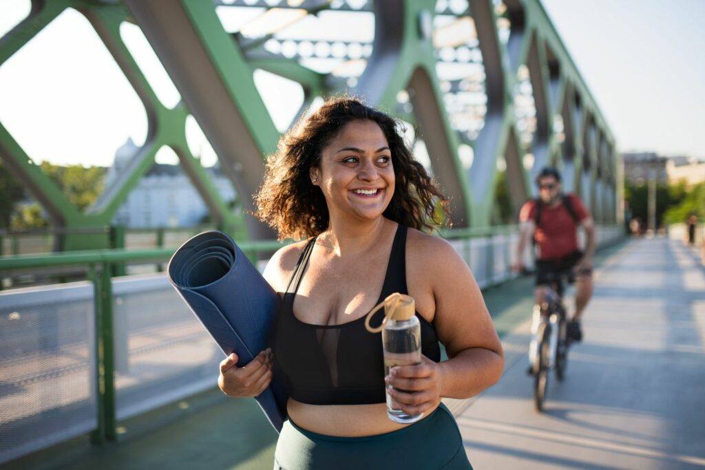 A woman walking outdoors with a yoga mat and water bottle.